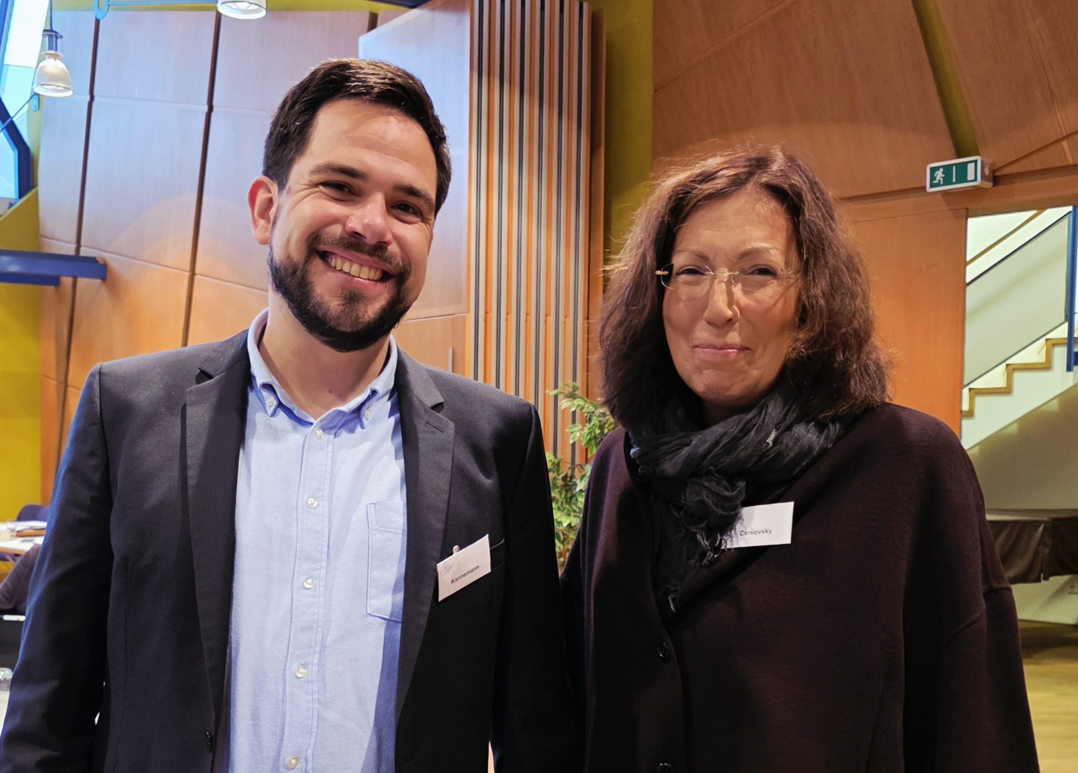 Ein jüngerer Mann mit Bart und eine Frau mit schulterlangen Haaren und randloser Brille lächeln in die Kamera, dahinter eine Treppe und vertäfelte Wände.