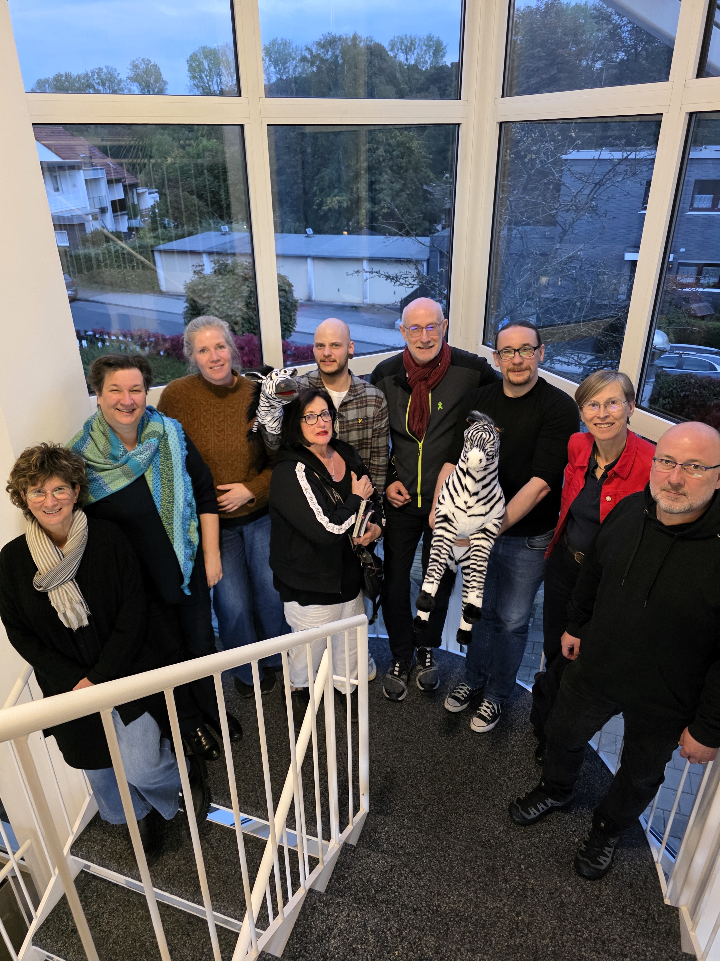 Neun Menschen stehen auf einer Treppe vor einem großen Fenster in der Abenddämmerung und lächeln nach oben in die Kamera. Ein großes Stoffzebra und eine Zebra-Handpuppe ergänzen das Gruppenbild. 