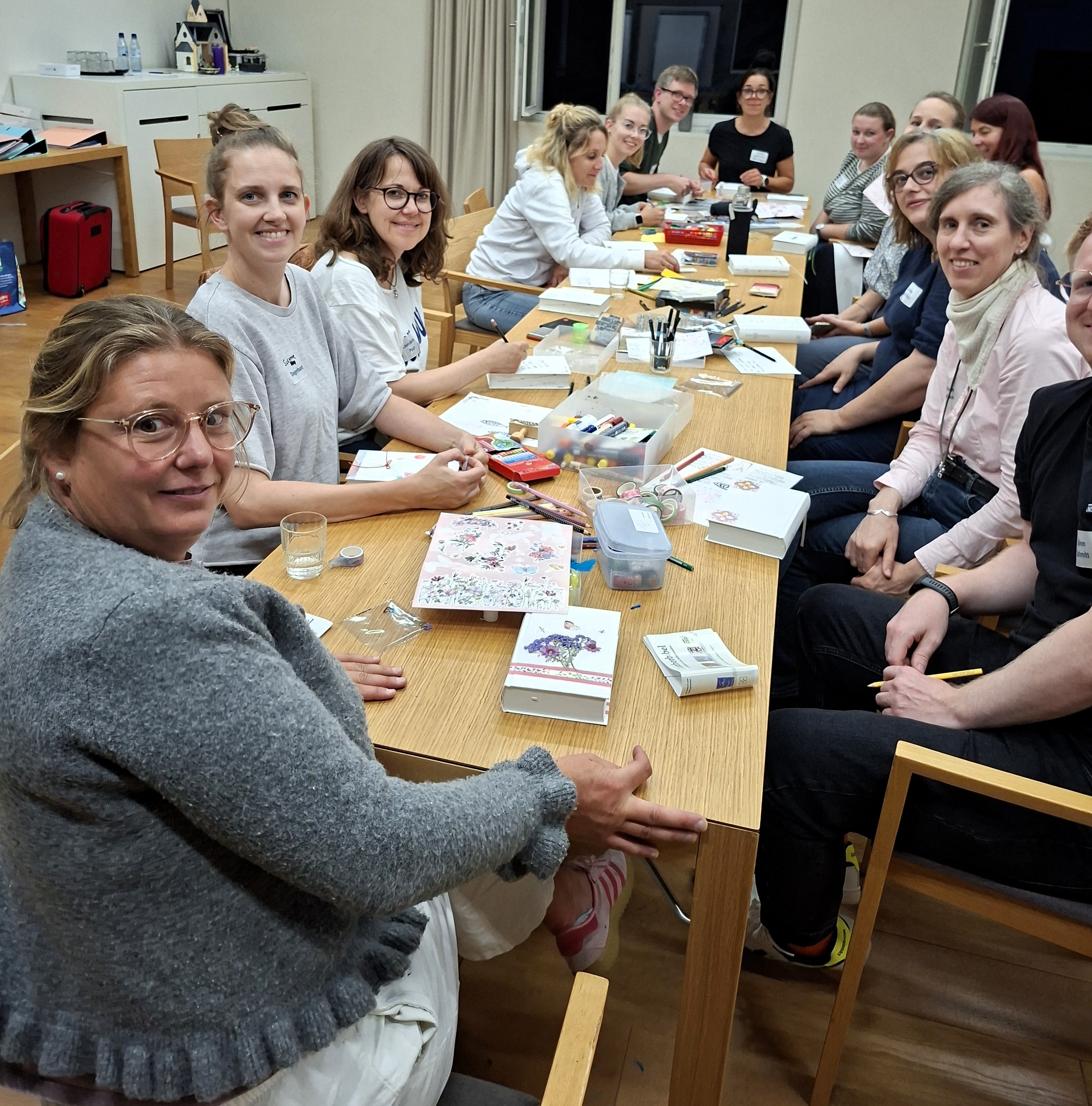 Menschen sitzen an einem Tisch und schauen in die Kamera. Auf dem Tisch dicke weiße Bücher und Stifte sowie Bastelmaterialien.  Stifte 