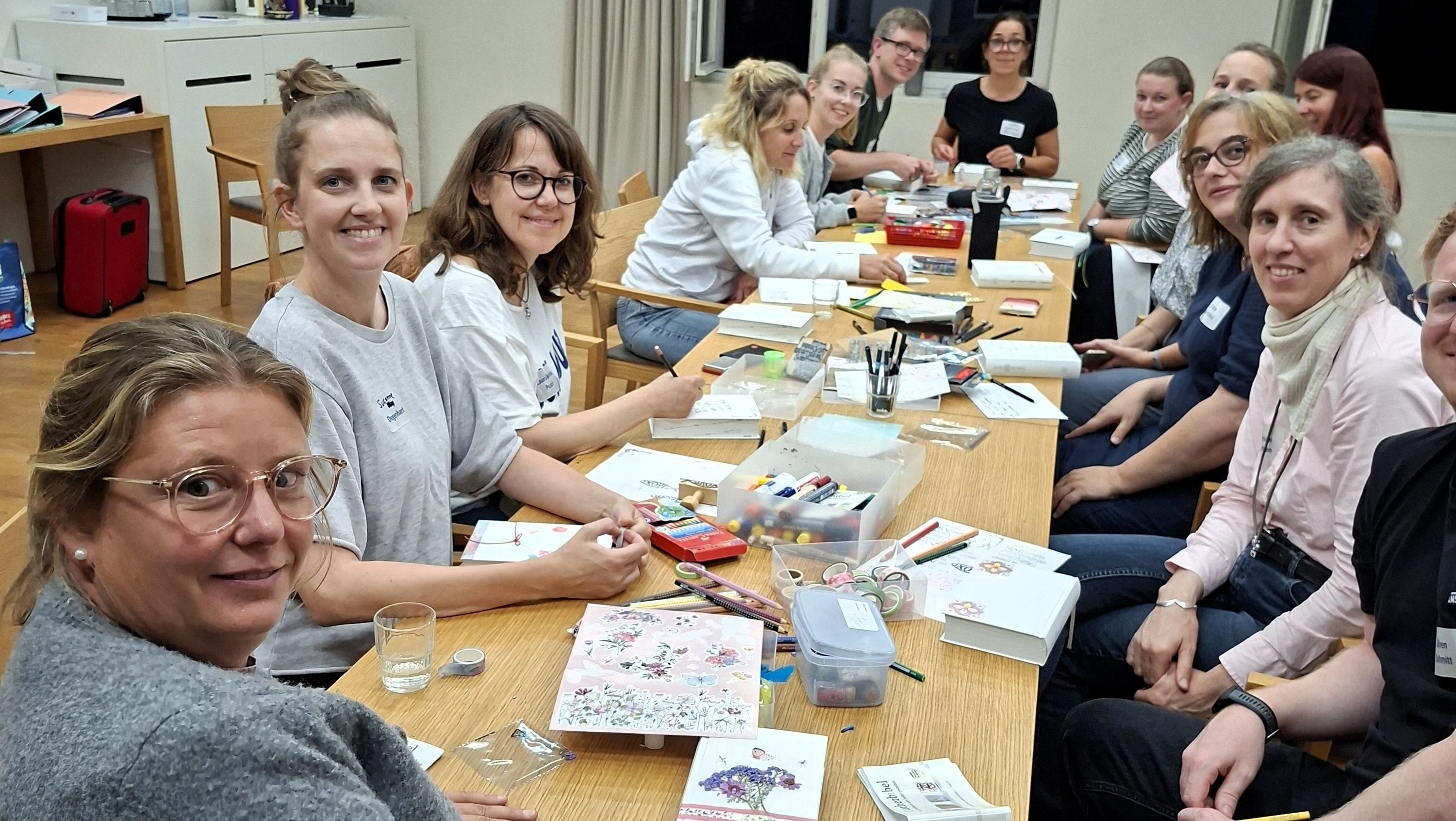 Menschen sitzen an einem Tisch und schauen in die Kamera. Auf dem Tisch dicke weiße Bücher und Stifte sowie Bastelmaterialien.  Stifte 